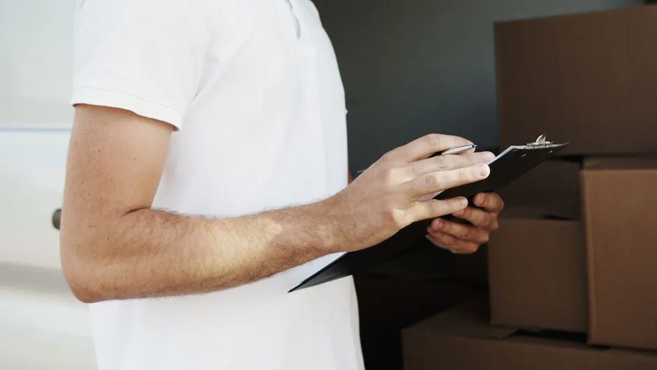 A delivery person in a white shirt holding a clipboard and pen near a vehicle with boxes.